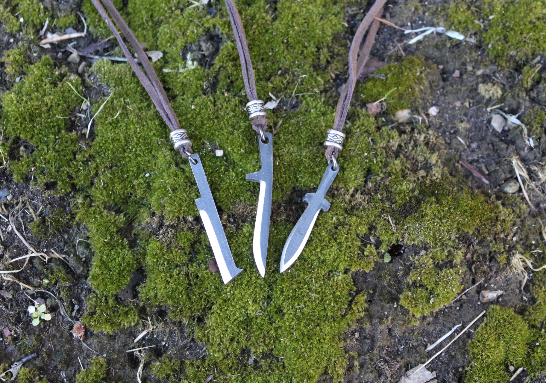 Three handmade knife and sword pendants on leather cords resting on moss